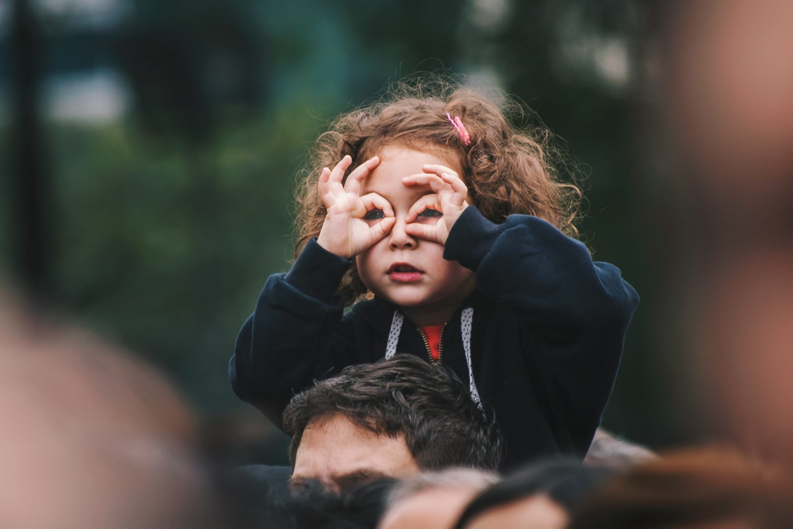 A photo of a small girl using her fingers as glasses, sat on a male relative's shoulders. Photo by Edi Libedinsky on Unsplash