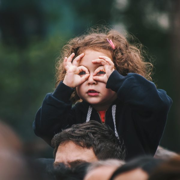 A photo of a small girl using her fingers as glasses, sat on a male relative's shoulders. Photo by Edi Libedinsky on Unsplash