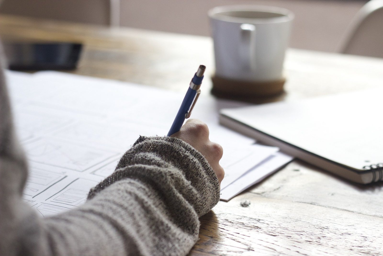 A person writing with a pen and paper- a coffee mug can be seen in the background.