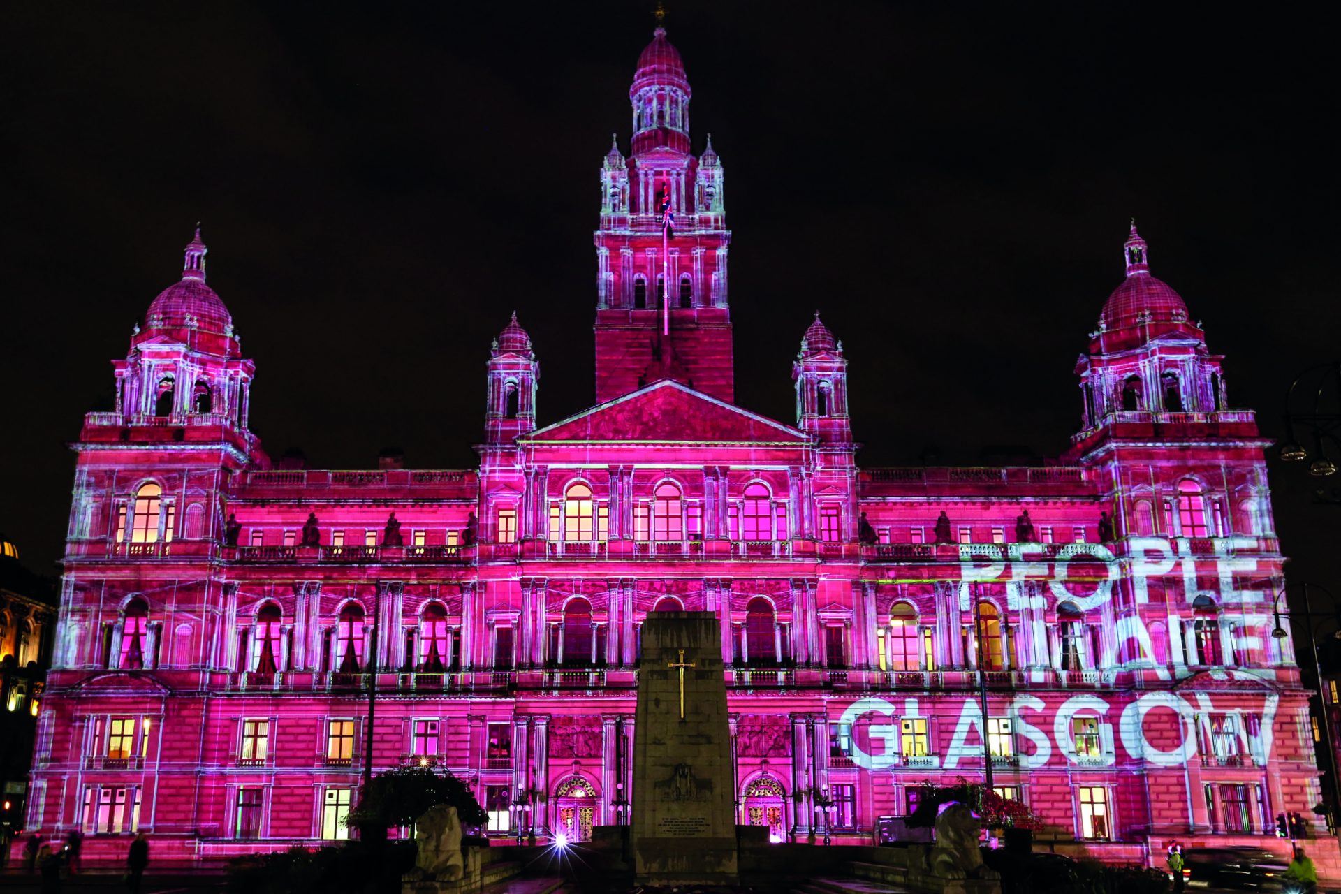 A picture of a building at night- it is lit up in pink with the words 'people make Glasgow' projected on the outside of the building.