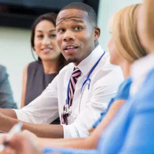 A diverse group of professionals sit around a meeting table engaged in discussion. A man in a shirt and tie is speaking and gesturing while colleagues listen attentively, creating a collaborative and inclusive workplace setting.