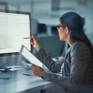 A person wearing glasses sits at a desk in an office, reviewing information on a large computer monitor while holding a tablet and pointing at the screen with a pen. The image represents focused computer-based work, data analysis, or document review.